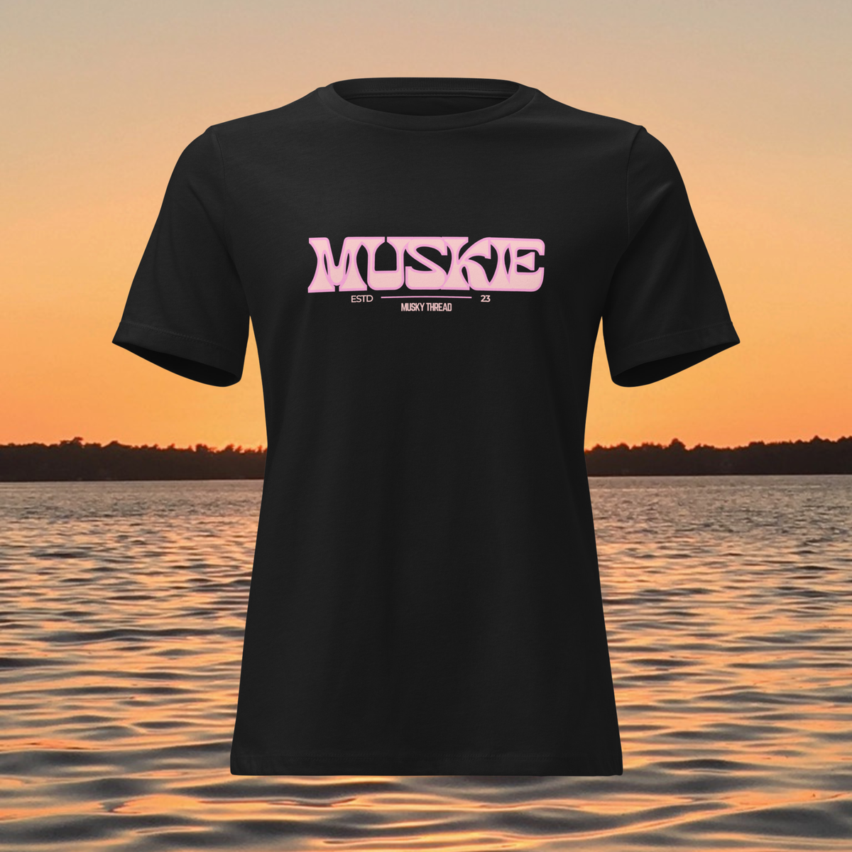 A black short-sleeve t-shirt with a pink graphic reading 'MUSKE ESTD 25 MENDT THREAD' on the chest, shown against a sunset over water background.