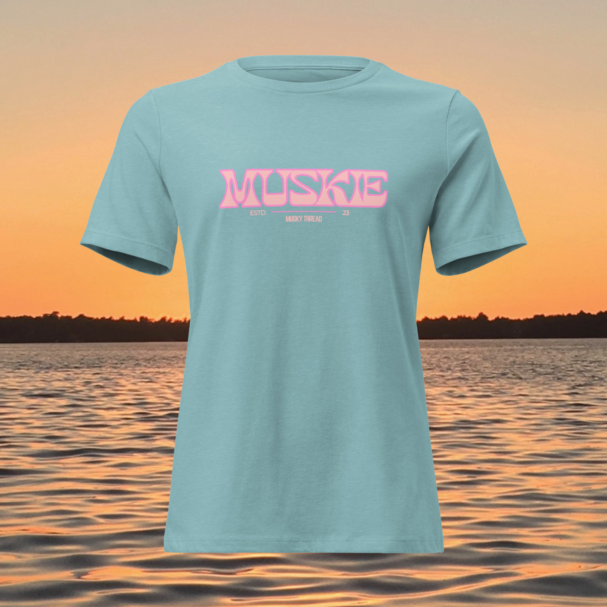 A light blue short-sleeve t-shirt with a pink 'MUSKE' graphic printed across the chest, displayed against a sunset lake background.