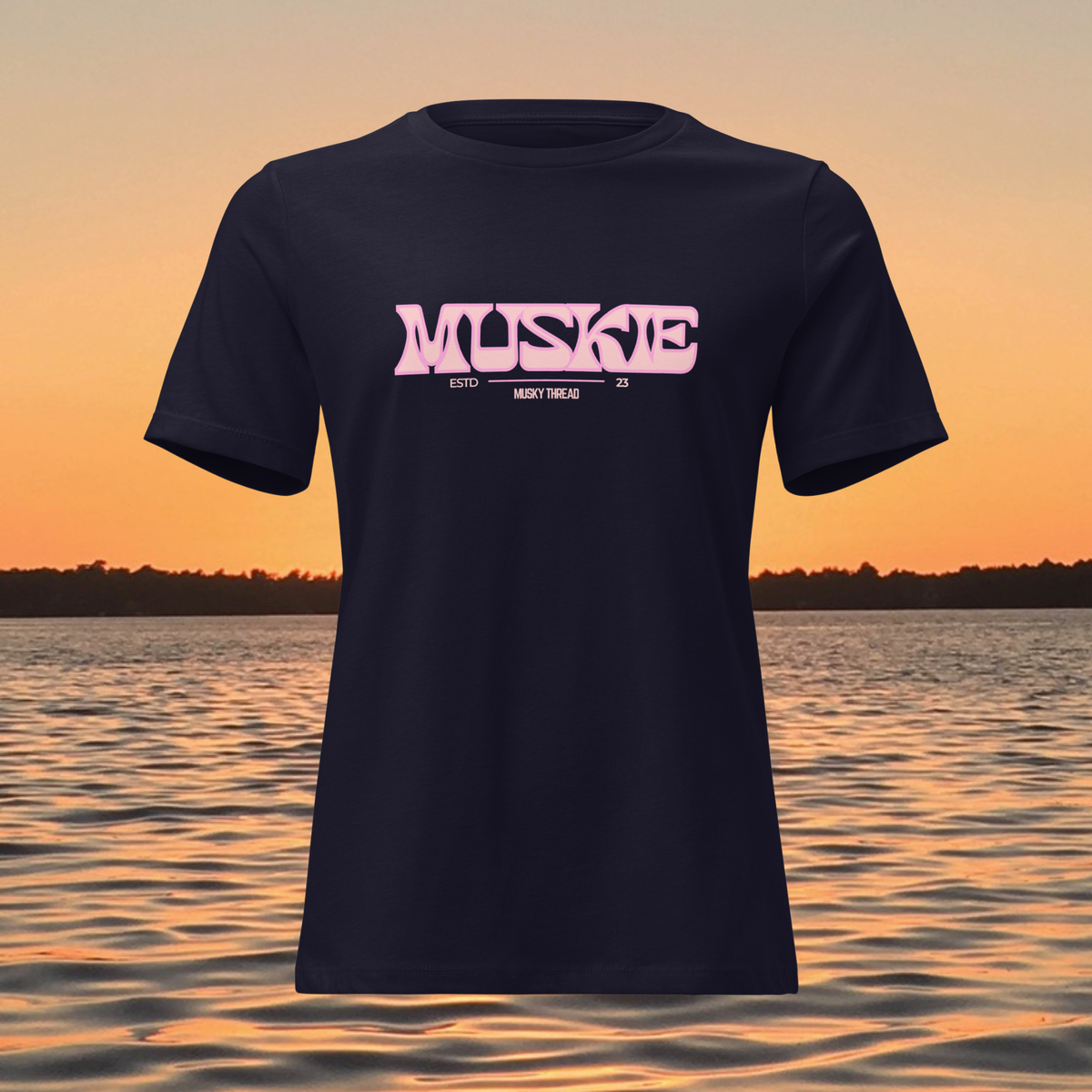 A navy blue short-sleeve t-shirt with a pink graphic reading 'MUSKE ESTD 25 MADE IN THROUGH' on the chest, shown against a sunset over water background.