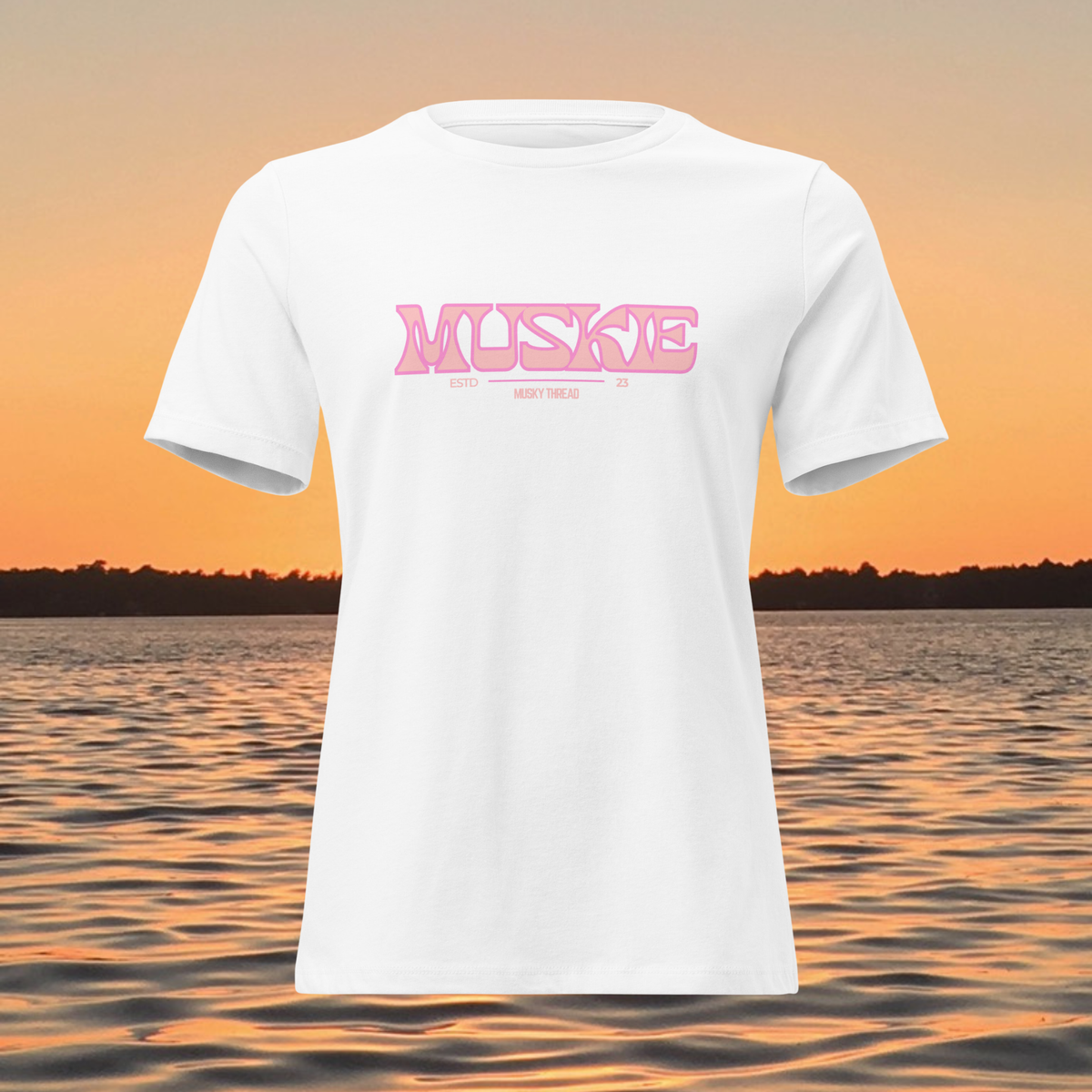 A white short-sleeve t-shirt with a pink 'MUSKE' graphic printed across the chest, displayed against a sunset lake background.