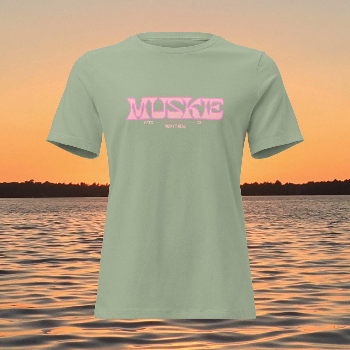 A sage green short-sleeve t-shirt with a pink retro-style 'MUSKE' graphic on the chest, displayed against a sunset lake background.