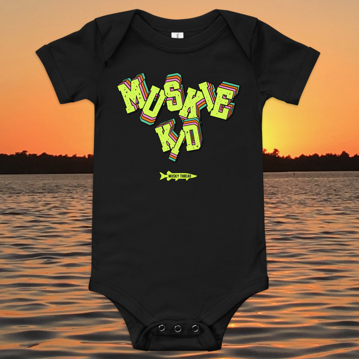 baby one-piece with neon green, pink, and teal "Muskie Kid" retro graphic and Musky Thread fish logo, shown against a lake sunset background